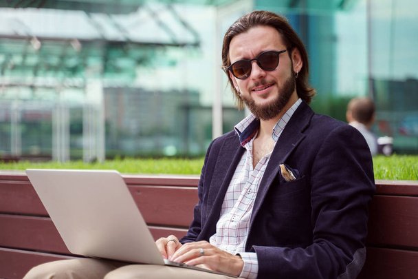 Photo of young brunet businessman with laptop sitting on wooden bench in city center on summer day - Fotoğraf, Görsel