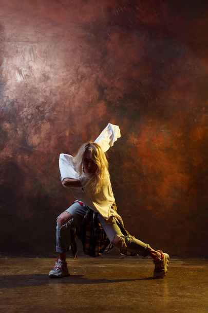 Photo of long-haired blonde woman looking at camera in ripped jeans dancing on brown background - Φωτογραφία, εικόνα