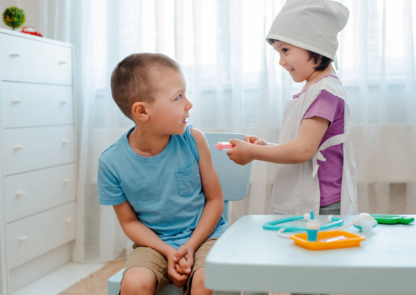 Дети играют в больнице. Девушка одета в врача 4 года делает инъекцию мальчику 6 лет с игрушечным шприцем в комнате в детском саду. - Фото, изображение