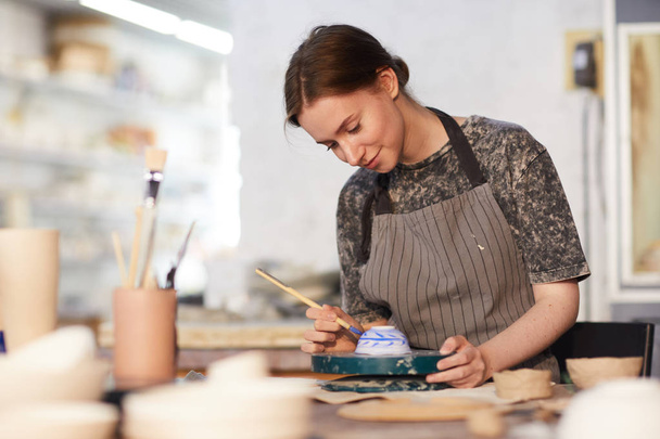 Inhoud aantrekkelijk geschoolde jonge dame in schort staan aan tafel en het tekenen van blauwe bladeren op keramische kom in aardewerk workshop - Foto, afbeelding
