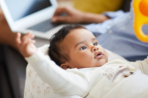 Primer plano de contenido curioso bebé niño en traje de cuerpo acostado en capullo y mirando sonajero mientras la madre trabaja con el ordenador portátil en el fondo
 - Foto, Imagen