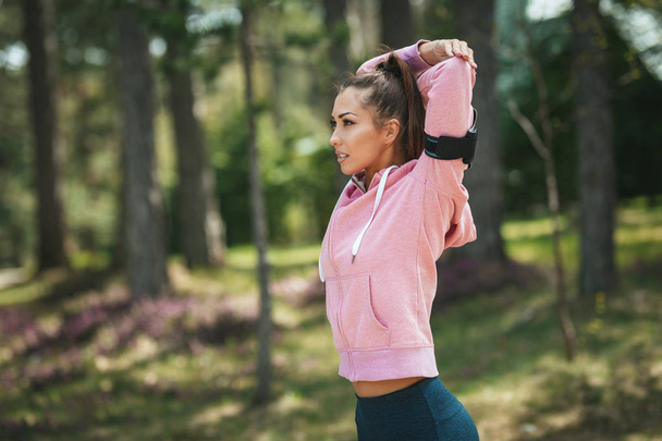 glückliche Frau in pastellfarbener Sportbekleidung, die Musik hört und sich im sonnigen Wald dehnt - Foto, Bild