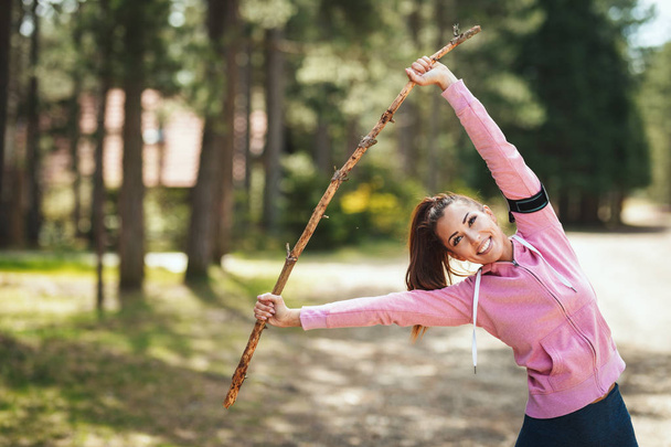 Happy woman with headphones stretching in a sunny forest trail, enjoying landscape among the trees. - Foto, Bild