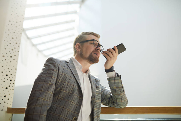 Уверенный в себе красивый бородатый мужчина в очках, опирающийся на перила и записывающий голосовое сообщение на смартфон
 - Фото, изображение
