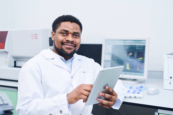 Zufriedene junge schwarze Medizinerin mit Bart und Schnurrbart sitzt am Schreibtisch mit modernem Computer und nutzt digitales Tablet im Labor - Foto, Bild