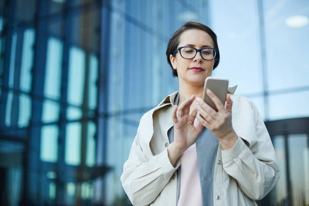 Serieuze drukke aantrekkelijke vrouw van middelbare leeftijd in vacht staande tegen kantoorgebouw en het gebruik van smartphone tijdens het controleren van e-mail - Foto, afbeelding
