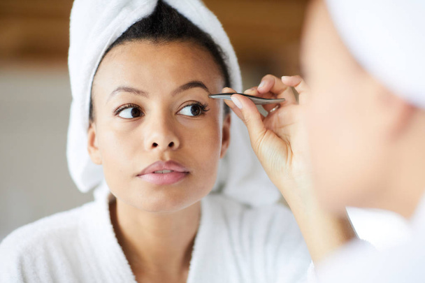 Head and shoulders portrait of  beautiful Mixed-Race woman plucking eyebrows looking in mirror during morning routine, copy space - Photo, Image