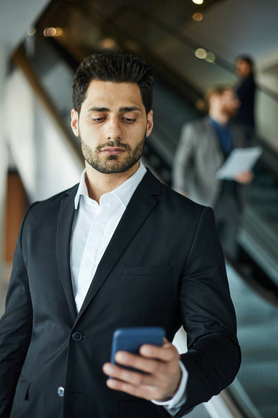 Seriózní pohledný mladý podnikatel se smíšenými závody v černém obleku, který stojí na letišti a používá telefon Smartphone, který odpovídá mobilním App - Fotografie, Obrázek