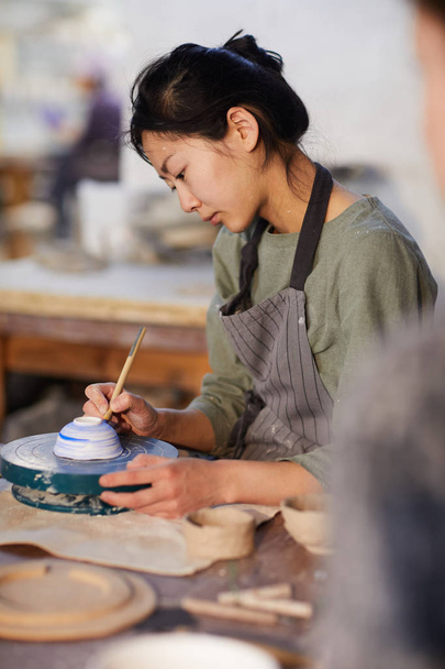Serieuze geconcentreerde jonge Aziatische vrouw in schort zittend aan tafel en schilderij keramische kom met penseel in workshop - Foto, afbeelding
