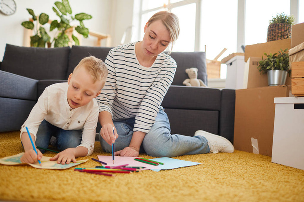 Sério mãe atraente e seu filho criativo sentado no tapete na sala de estar com caixas móveis e desenho juntos
 - Foto, Imagem