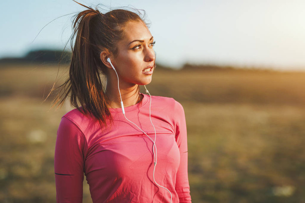 Nuori nainen vaaleanpunaisissa urheiluvaatteissa kuuntelemassa musiikkia ja valmistautumassa lenkkeilyyn aamulla vuorilla
 - Valokuva, kuva