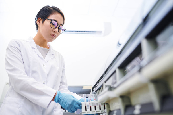 Grave atraente jovem funcionário do laboratório asiático em óculos vestindo jaleco e luvas cirúrgicas colocando amostras de sangue na máquina
 - Foto, Imagem