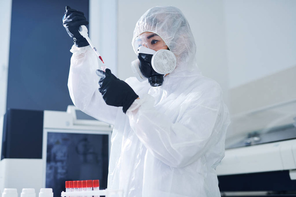 Científico sanitario concentrado serio en traje de riesgo biológico y respirador de pie en el laboratorio y dejando caer la sangre mientras se analiza la muestra de virus
 - Foto, imagen
