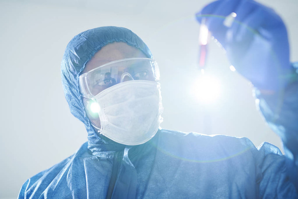 Concentrated black infectious disease scientist in biohazard suit and mask holding test tube and analyzing virus-infected blood sample - Photo, Image