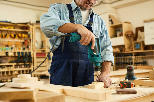 Porträt eines reifen Tischlers, der Holz bohrt, während er in der Tischlerei arbeitet, Kopierraum - Foto, Bild