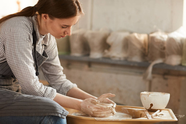 Poważna Ładna młoda kobieta z kucyk siedzi na kole garncarstwa i dotykając gliny z rękami podczas tworzenia garnka ceramicznego w warsztacie - Zdjęcie, obraz
