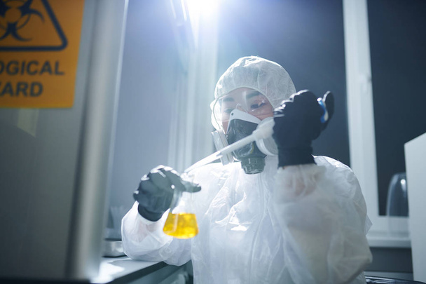 Microbiólogo concentrado con gafas protectoras, traje de riesgo biológico y máscara respiratoria que deja caer el reactivo en el frasco mientras estudia la sustancia en el laboratorio
 - Foto, Imagen