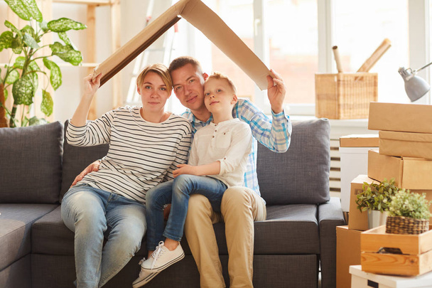 Portrait of content young family in casual clothing sitting on gray sofa and holding cardboard roof above their heads - Foto, imagen