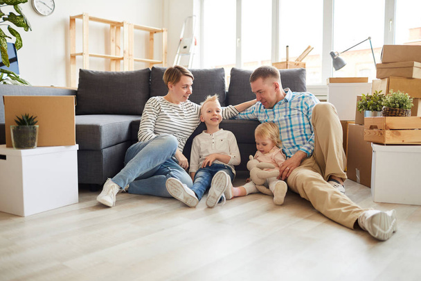 Csoport pozitív fiatal család alkalmi ruhák ül a padlón, és támaszkodva a kanapén, miközben egyre telepedett le az új otthon - Fotó, kép