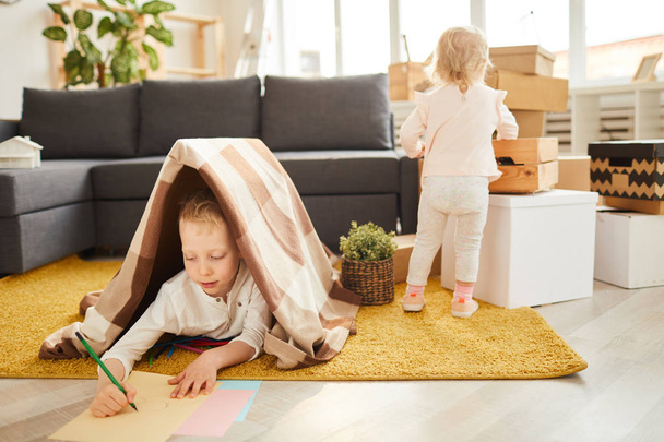 Children getting settled in new apartment: concentrated little boy lying under plaid on carpet and drawing picture while his younger sister unpacking boxes - 写真・画像