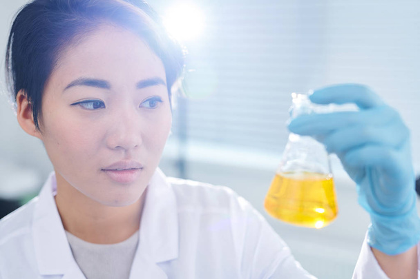 Un jeune ouvrier de laboratoire asiatique sérieusement perplexe portant des gants de caoutchouc debout au soleil et examinant un liquide chimique dans une fiole
 - Photo, image