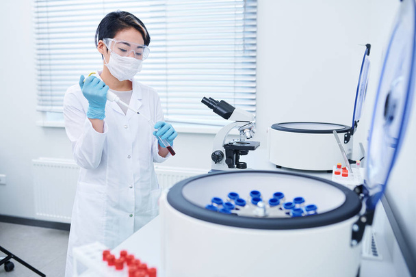 ernsthafter, konzentrierter junger asiatischer Laborant mit kurzen Haaren, weißem Mantel, Maske und Schutzbrille, der am Arbeitsplatz Reagenzien mit Blutproben mischt - Foto, Bild