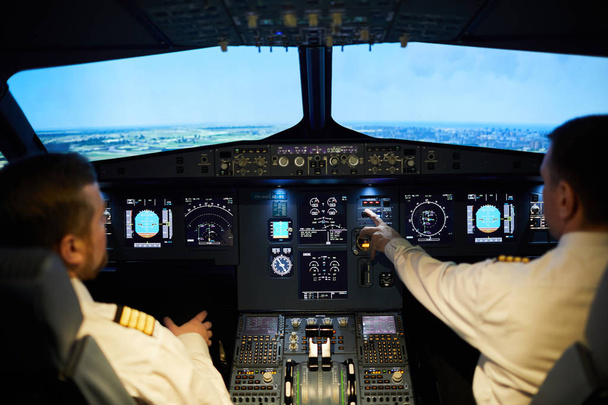 Zespół profesjonalnych pilotów siedzących na panelu sterowania z różnymi przełącznikami i uchwytami oraz latającym samolotem razem w niebie - Zdjęcie, obraz