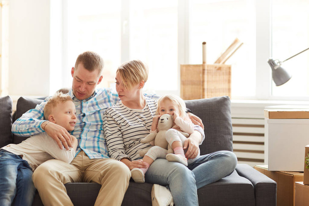 Positive beautiful young family in casual clothing sitting on comfortable sofa and embracing each other while chatting together at home - Zdjęcie, obraz