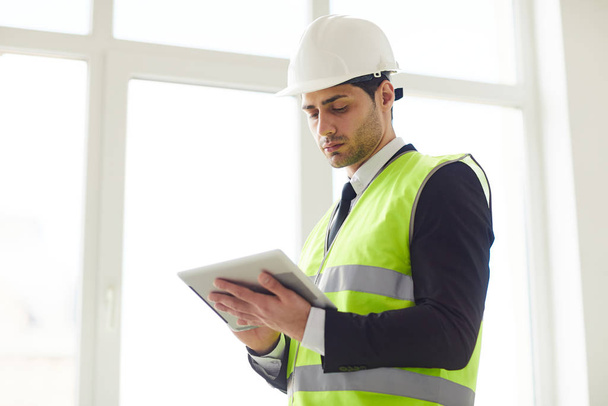 Waist up portrait of  Middle-Eastern engineer wearing hardhat standing against window  holding tablet, copy space - Photo, Image