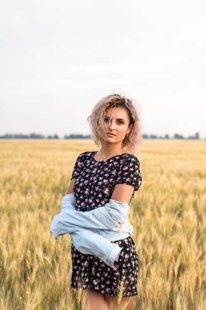 young tanned curly haired woman in denim jacket and dress on the field of ripened wheat, sunset time - Photo, Image