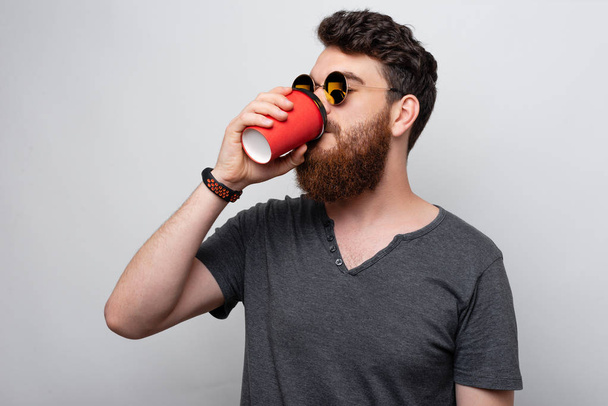 Knappe bebaarde jonge man die koffie of thee drinkt om in een papieren rode beker te gaan. - Foto, afbeelding