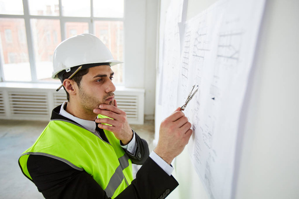 Retrato de vista lateral del pensativo ingeniero de Oriente Medio sosteniendo planes contra la pared mientras está de pie en el sitio de construcción
 - Foto, imagen