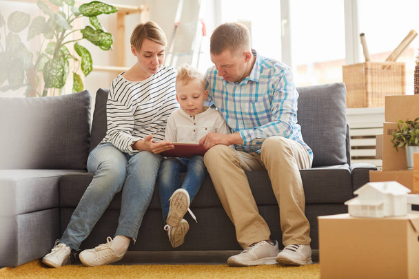 Seriózní zamyšlený mladí rodiče sedí kolem syna na pohovce a pomáhají synovi dělat domácí úkoly online pomocí moderního tabletu - Fotografie, Obrázek