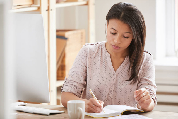 Portrait of beautiful Mixed race businesswoman writing in notebook while sitting at desk in office, copy space - Foto, Imagem