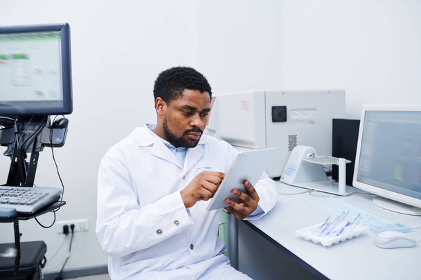 ernste konzentrierte junge schwarze Medizinerin mit Bart sitzt am Schreibtisch und nutzt digitales Tablet bei der Analyse von Daten im Labor - Foto, Bild