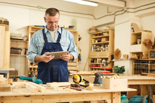 Retrato de cintura hacia arriba del carpintero senior usando tableta mientras trabaja en carpintería, espacio de copia
 - Foto, imagen