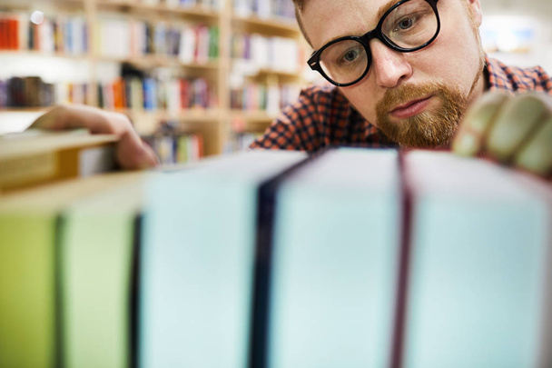 Primer plano de bibliotecario barbudo concentrado serio en gafas negras de pie en la estantería y encontrar libro
 - Foto, Imagen