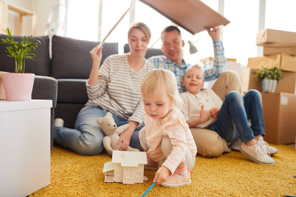 Serious cute little girl with blond hair sitting on carpet and playing with toy house while her parents and brother hiding under cardboard roof in background. - 写真・画像