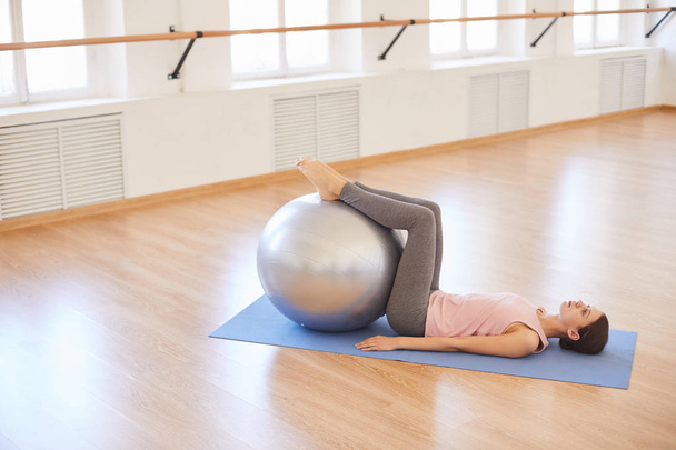 Kalm ontspannen jonge vrouw liggend op mat en het houden van voeten op Swiss ball terwijl het doen van oefening in lege Pilates Studio - Foto, afbeelding