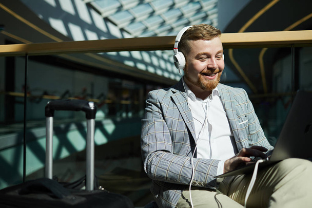 Cheerful excited chubby young man with beard sitting in waiting area of airport and using laptop while browsing internet and listening to music in headphones - Foto, Bild