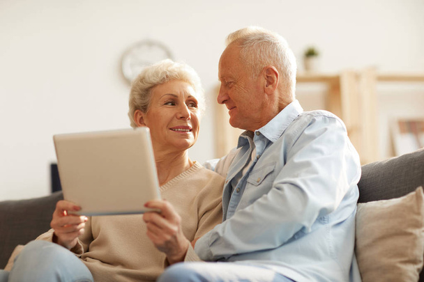 Низкоугловой портрет современной пожилой пары с помощью цифрового планшета дома, освещенный солнечным светом, копировальное пространство
 - Фото, изображение