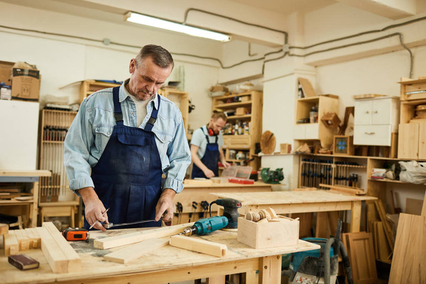 Porträt eines gestandenen Tischlers, der mit Holz arbeitet, das am Tisch in der Werkstatt steht, Kopierraum - Foto, Bild