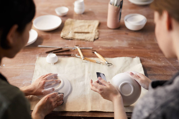 Rückansicht von nicht wiederzuerkennenden Handwerkerinnen, die an alten Holztischen sitzen und in der Werkstatt Tongeschirr mit Schleifpapier polieren - Foto, Bild