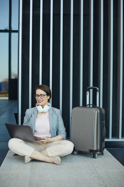 Positive attractive businesswoman with wired headphones on neck sitting with crossed legs on floor and working with laptop while waiting for boarding plane - Foto, afbeelding