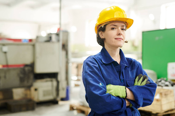 Μέση επάνω πορτρέτο του γυναίκα εργαζόμενη με αυτοπεποίθηση φορώντας σκληρό καπέλο κοιτάζοντας την κάμερα, ενώ ποζάρει στο εργοστάσιο, αντιγραφή χώρου - Φωτογραφία, εικόνα