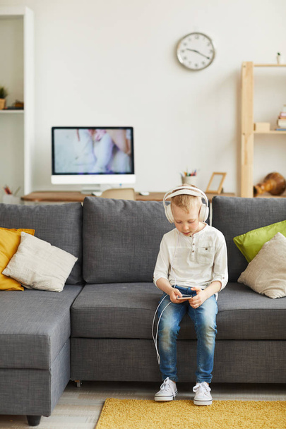 Vakava hipster pieni poika langallisissa kuulokkeissa istuu sohvalla olohuoneessa ja ladata musiikkia puhelimessa
 - Valokuva, kuva