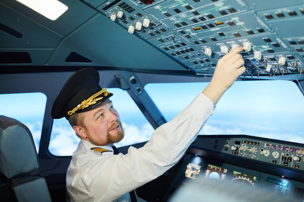 Sério gordinho barbudo piloto profissional em capitães cap sentado em poltrona e usando interruptores no cockpit enquanto voa avião
 - Foto, Imagem