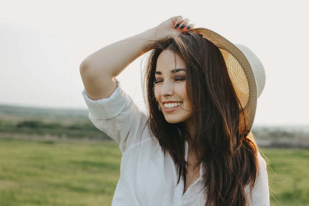 Portrait of charming carefree long hair asian girl in white clothes and straw hat enjoys life in field at sunset. Sensitivity to nature concept - Photo, Image