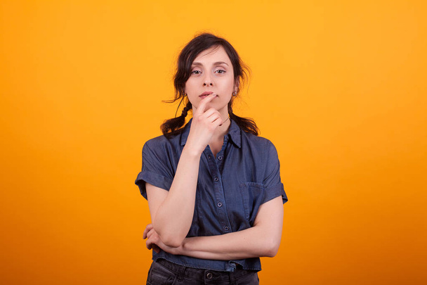 Thoughtful young woman in studio with hand near the face over yellow background - Foto, Imagen