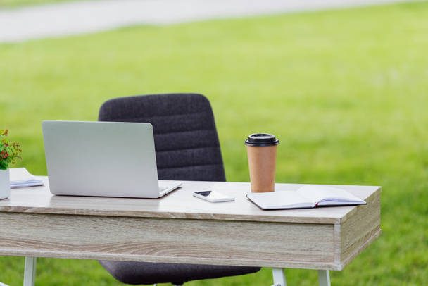 Bürotisch mit Laptop, Coffee to go, Notebook und Smartphone in der Nähe des Bürostuhls im Park - Foto, Bild